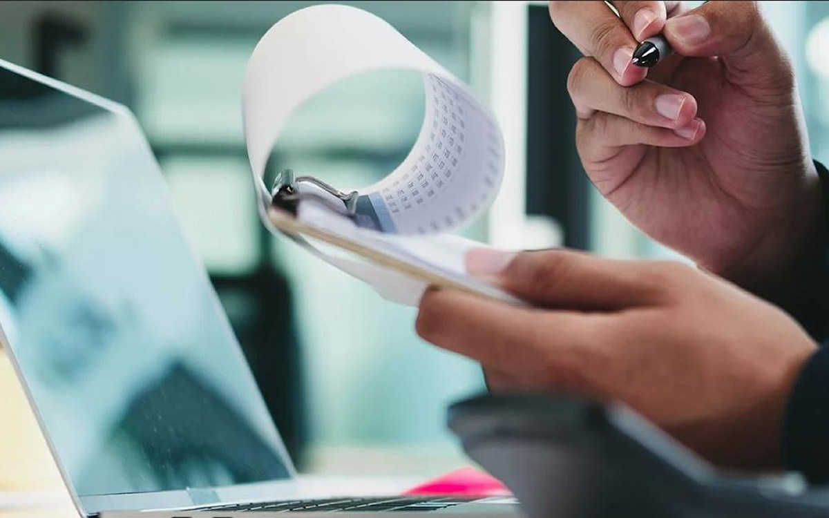 Person macht sich Notizen, daneben steht ein Laptop.